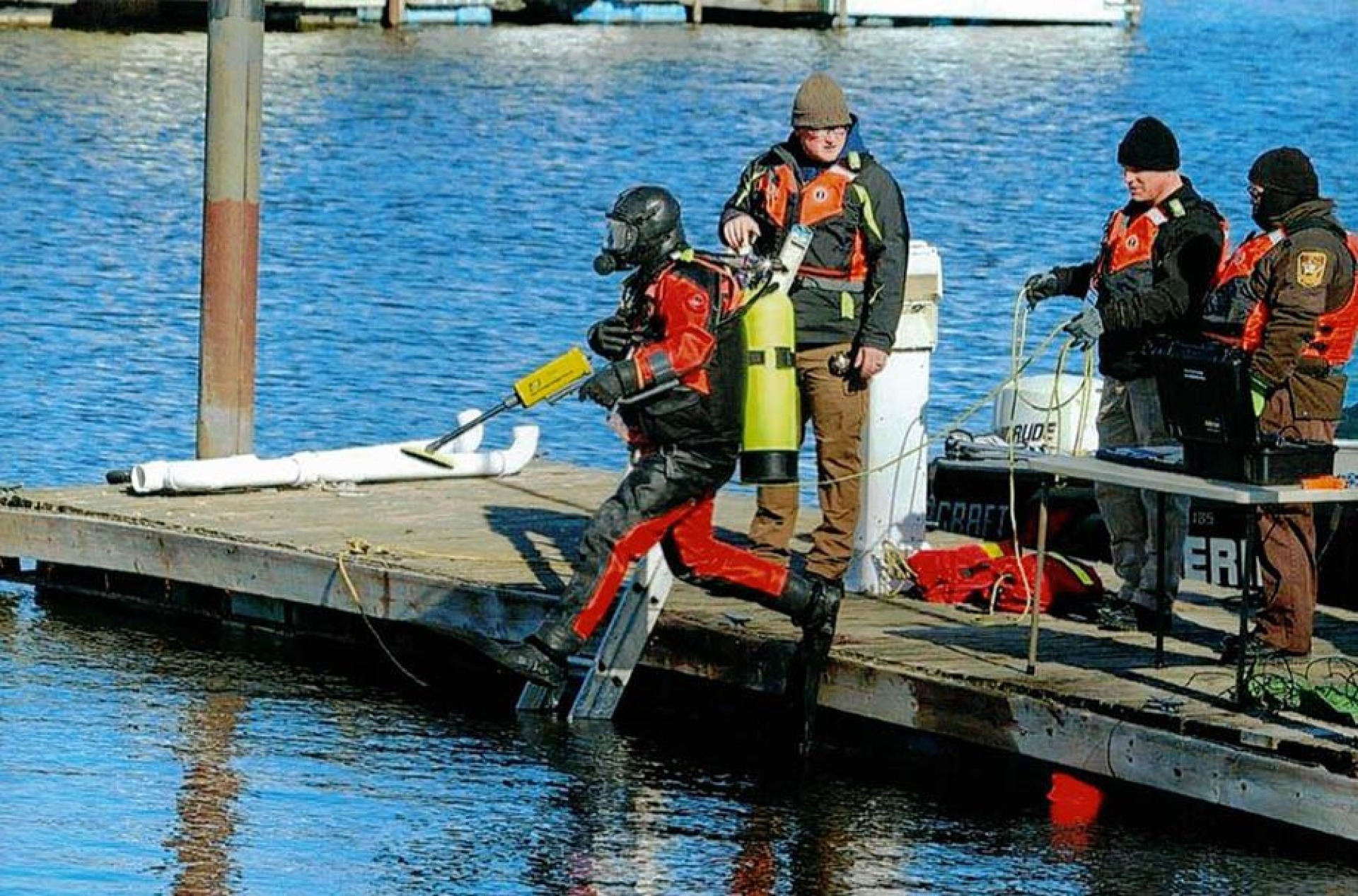 A dive/resue team member jumping off a dock into water with dive equipment on while others observe.